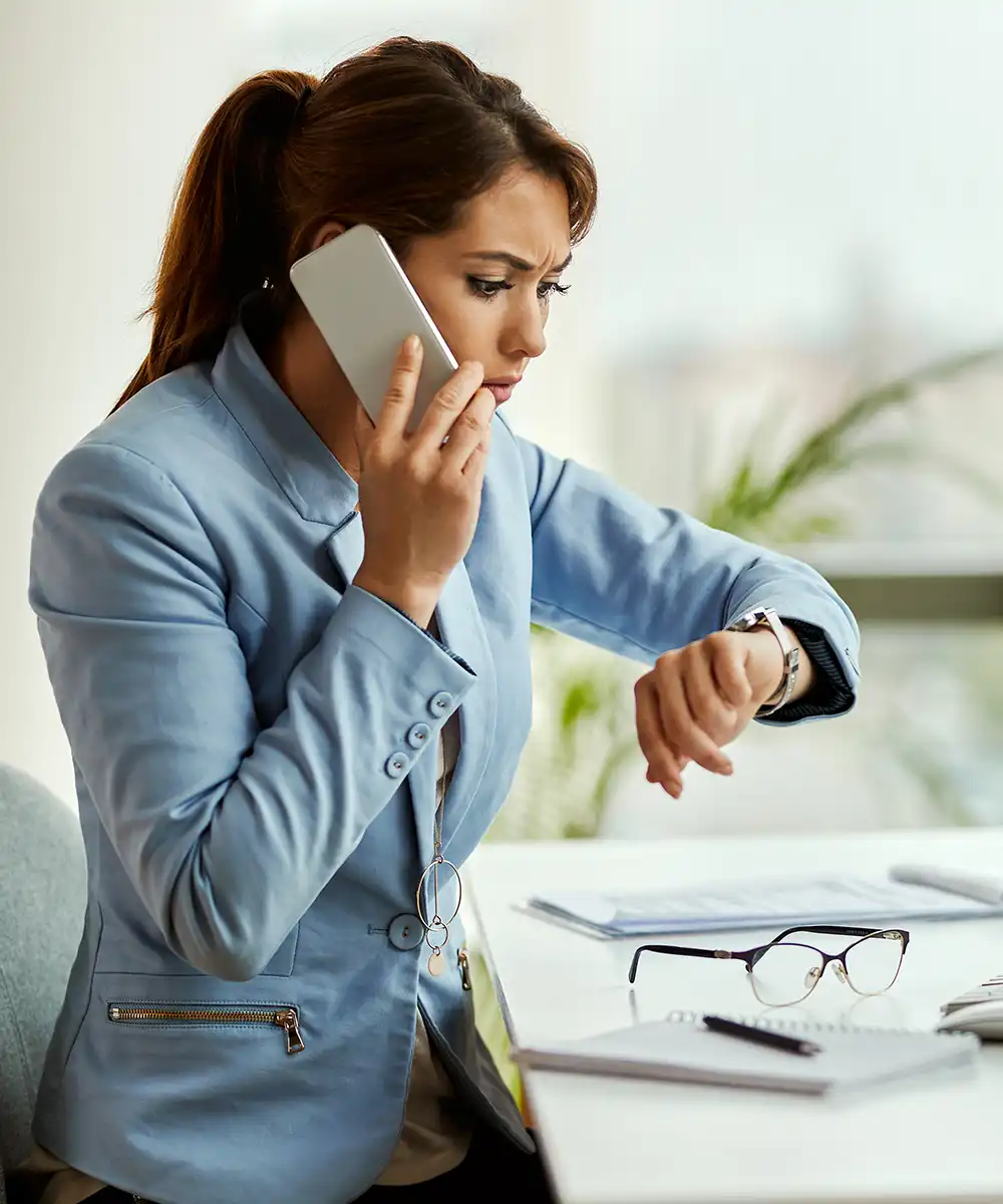 Mujer profesional hablando por teléfono mientras consulta su reloj en una oficina, mostrando preocupación y estrés por la falta de tiempo. Imagen que representa la presión laboral, la ansiedad, la sobrecarga de responsabilidades y la importancia del equilibrio emocional.